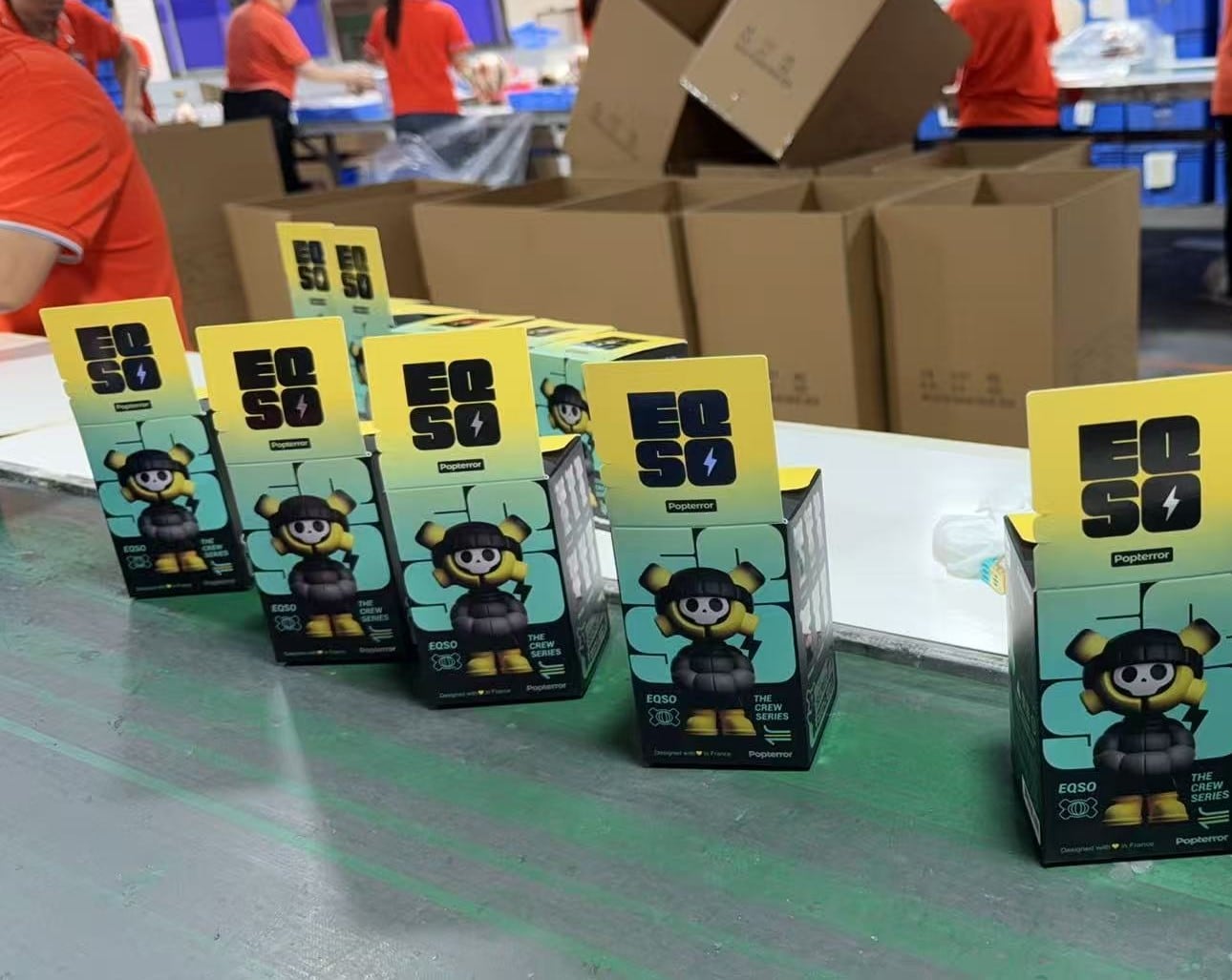 EQSO The Crew blind boxes lined up on a packing table, in front of shipping cartons, in our partner factory